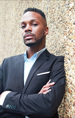 Dorian Issac wearing a black suit stands against a stone wall